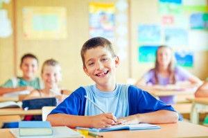 A smiling boy sits at a classroom desk, surrounded by books and school supplies