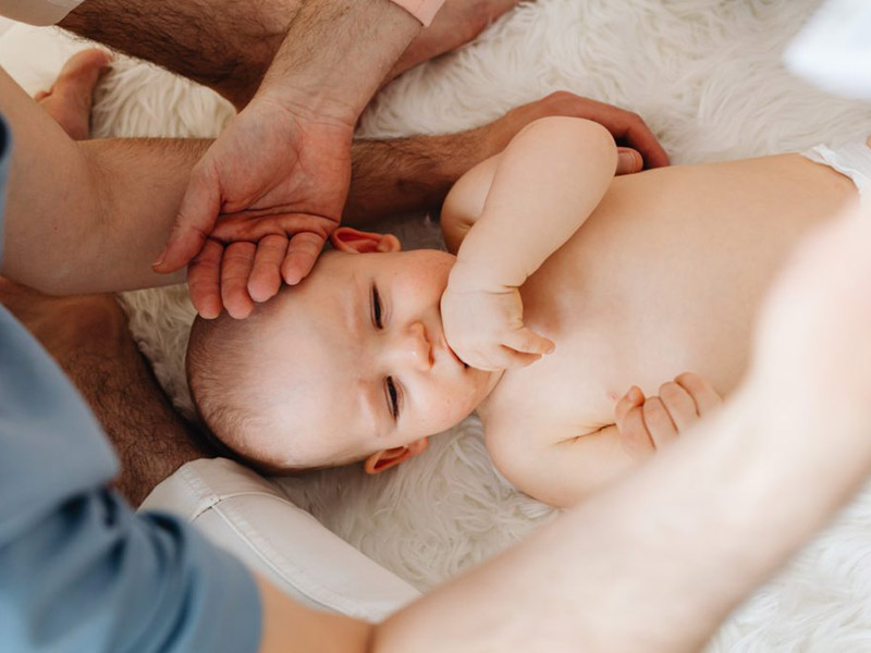 Baby sucking its thumb surrounded by two adults.