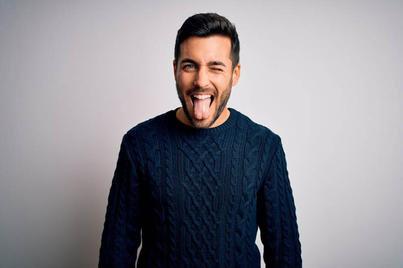 Young handsome man with beard wearing casual sweater standing over white background sticking tongue out happy with funny expression. Emotion concept.